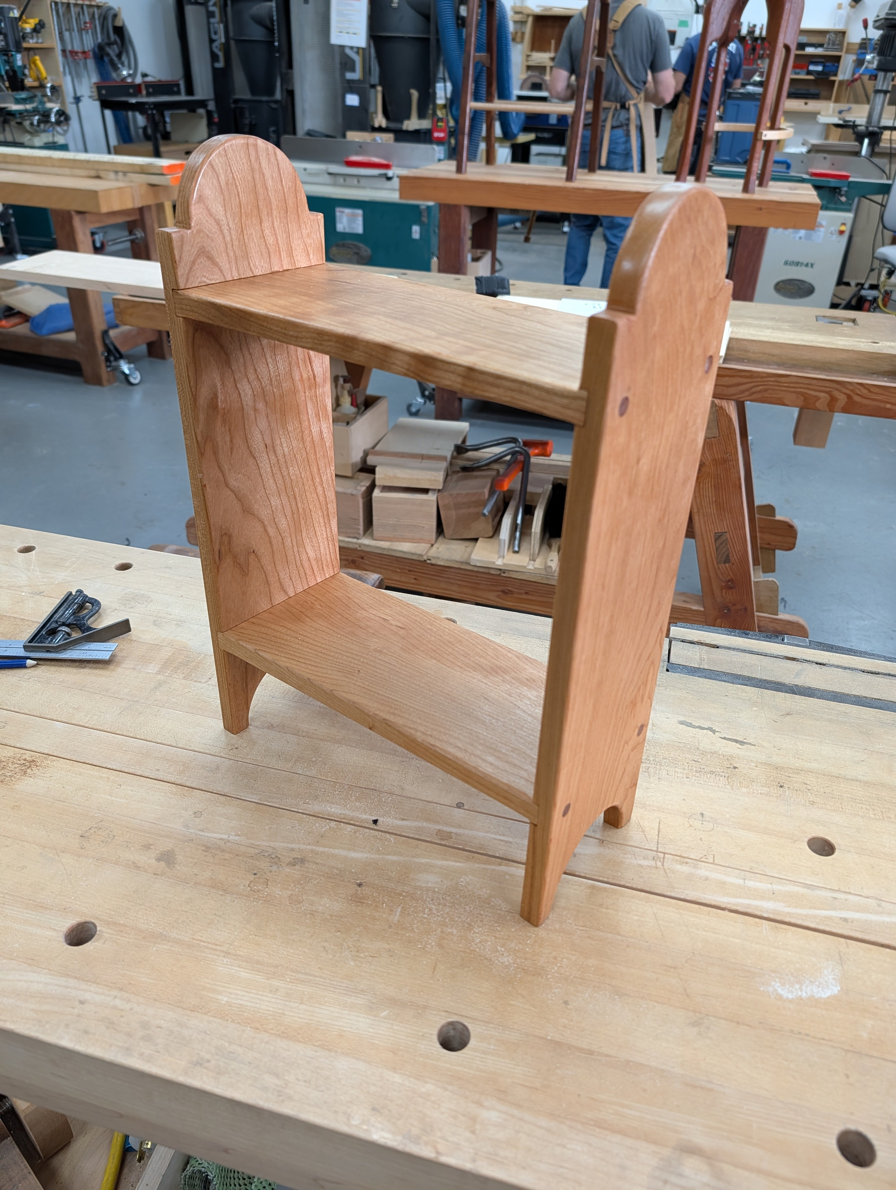 Auto-generated description: A small, handcrafted wooden shelf with two tiers is placed on a workbench in a woodworking shop.