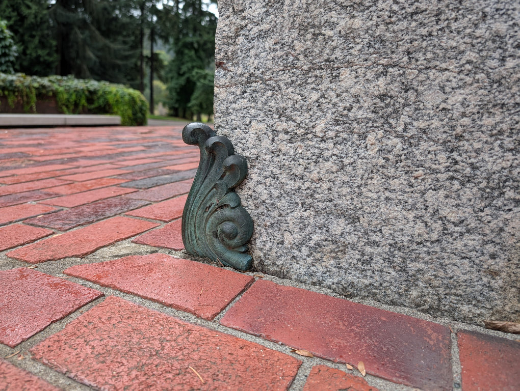 Auto-generated description: A decorative metal flourish is installed at the base of a stone wall where it meets a red brick pavement in an outdoor setting.