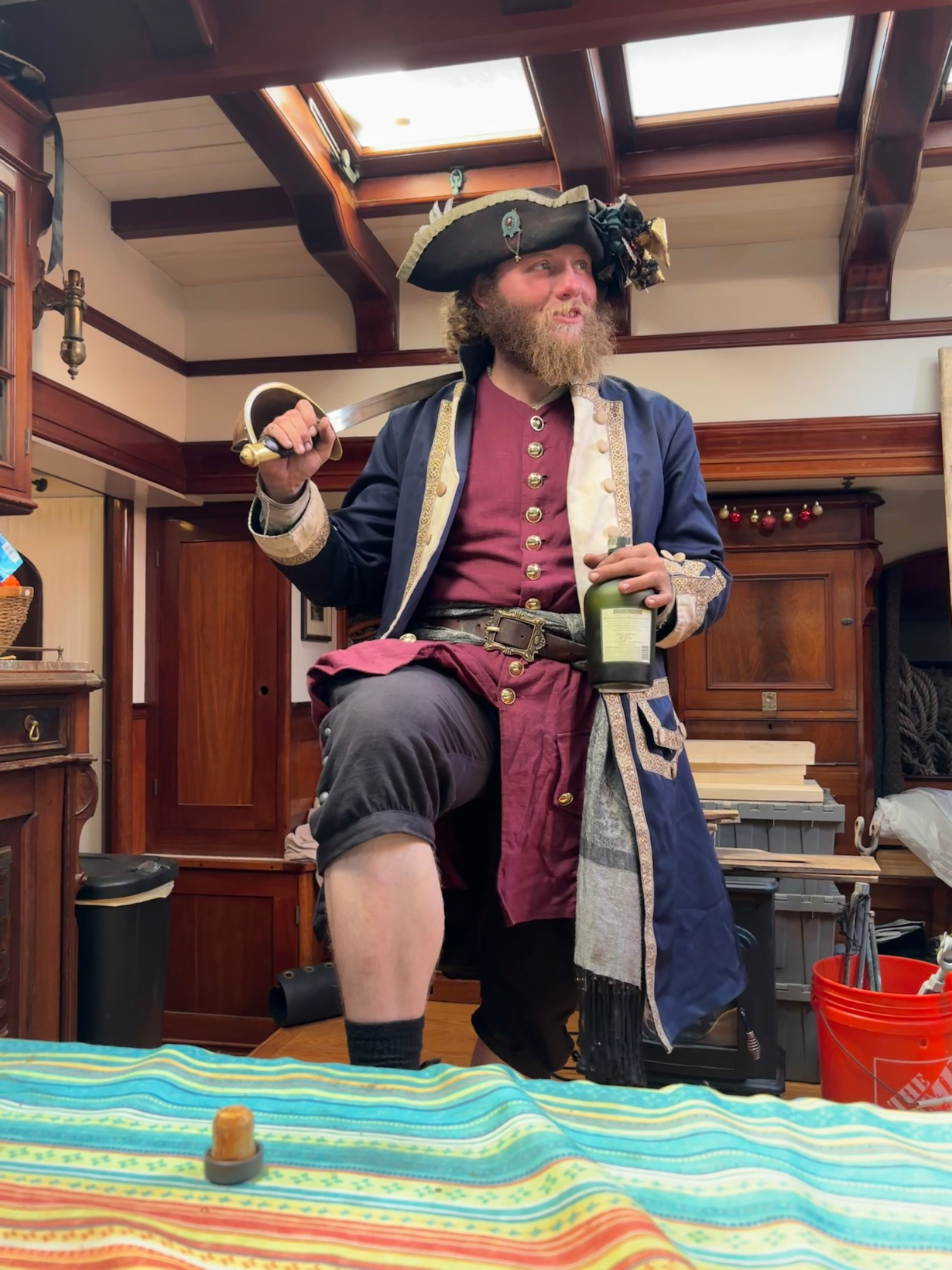 Auto-generated description: A person dressed as a pirate holding a sword and a bottle, standing inside a wooden room with a colorful tablecloth in the foreground.