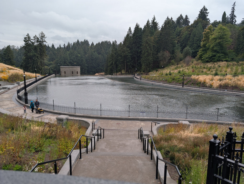 Auto-generated description: A calm, rectangular pond surrounded by a paved walkway and dense forest is viewed from the top of a staircase on a cloudy day.