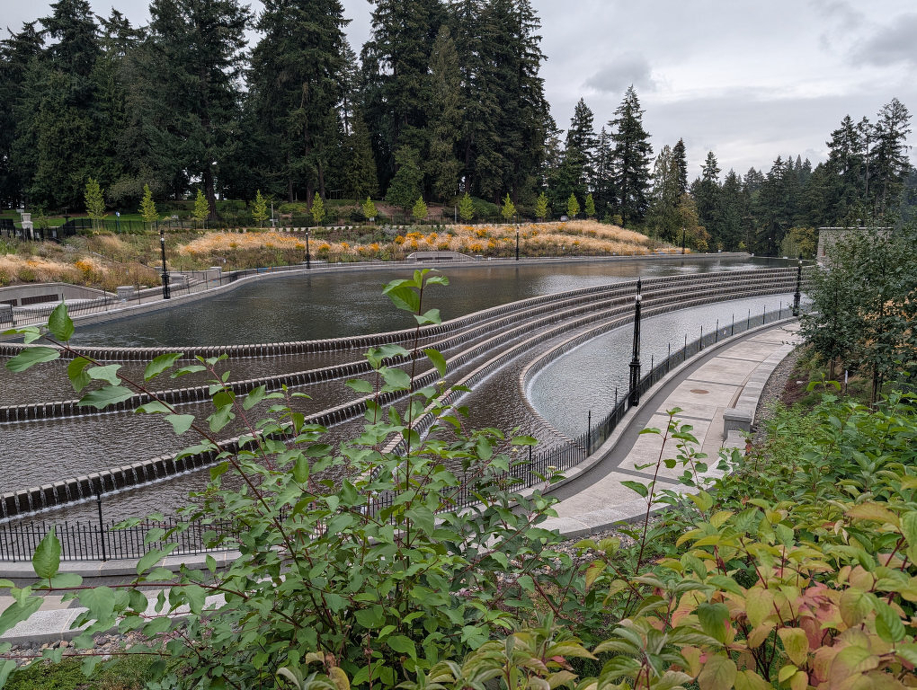 Auto-generated description: A curved, multi-tiered outdoor reservoir surrounded by greenery and tall trees under a cloudy sky.