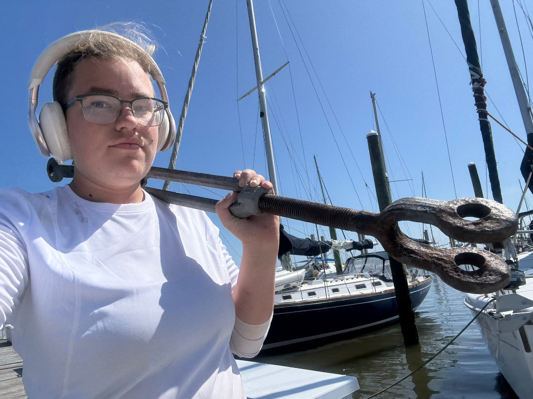 Auto-generated description: A person wearing headphones holds a large metal tool near a marina with sailboats in the background under a clear blue sky.