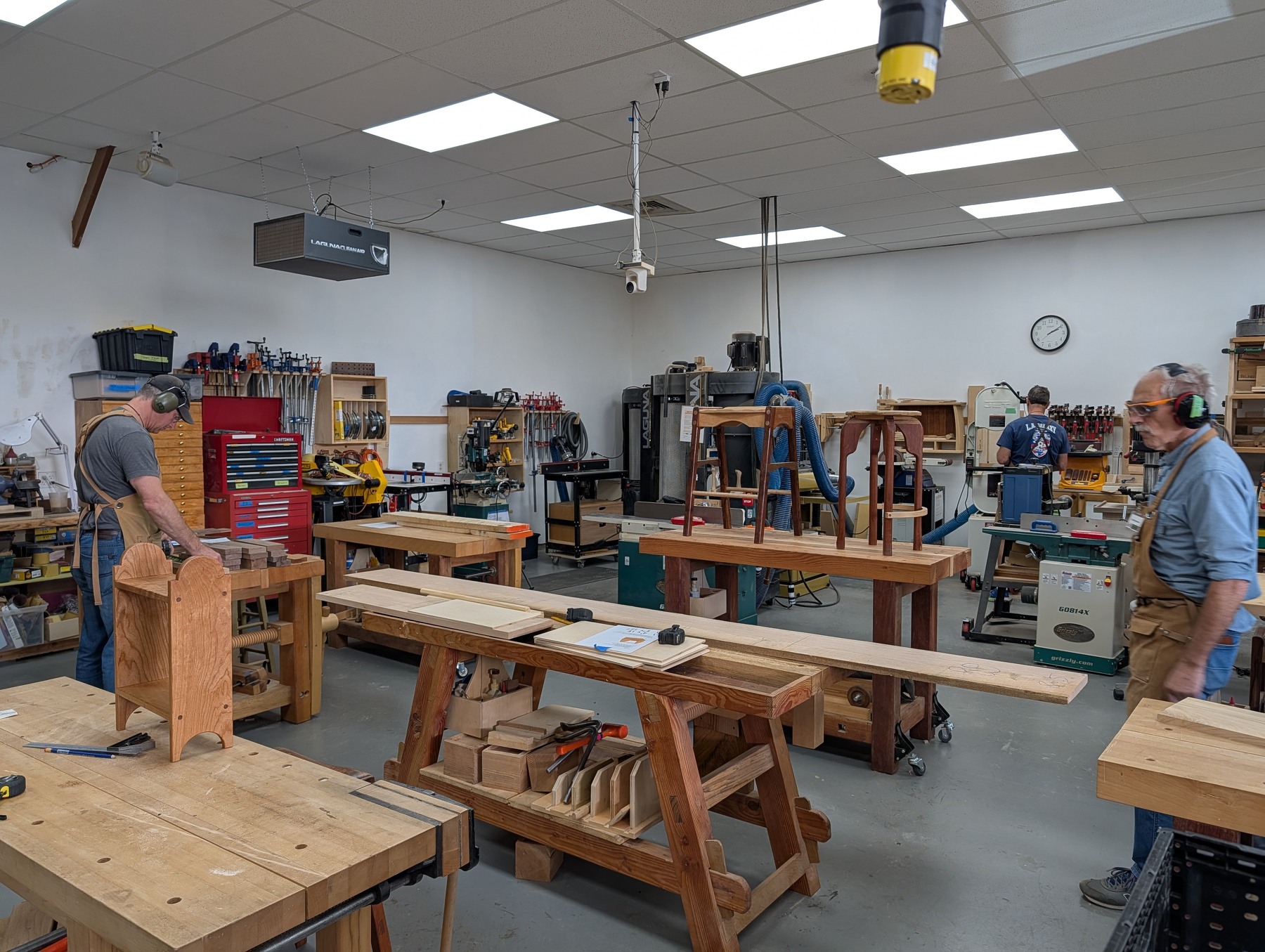 Auto-generated description: A spacious woodworking workshop with several people working on various wood projects at large workbenches.
